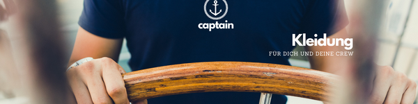 Junger Mann in dunkelblauem T-Shirt, steht an einem Steuerrad aus Holz. Das dunkelblaue Shirt ist bedruckt mit einem kleinen Anker Motiv und dem Schriftzug Captain. Eine Verlinkung auf diesem Bild führt direkt zur Kategorie Kleidung.